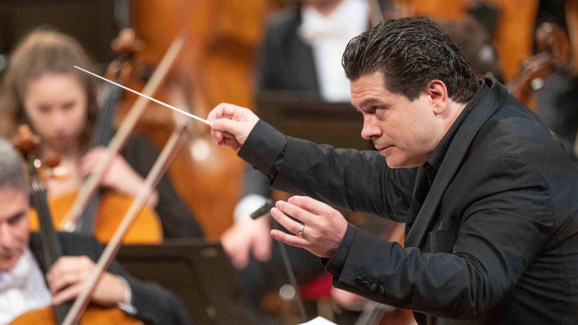 Cristian Măcelaru schaut konzentriert, hinter ihm musizieren Mitglieder des Orchesters.