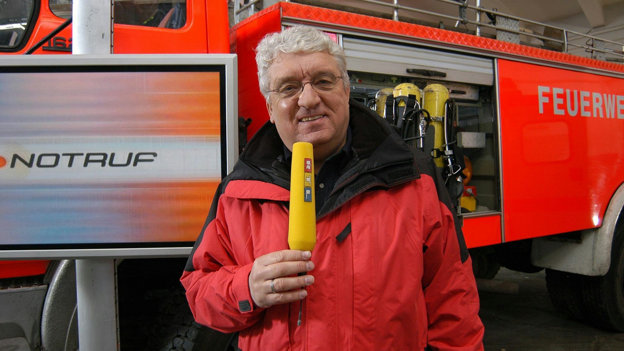 Moderator Hans Meiser steht vor einem Feuerwehrauto. Er trägt eine rote Jacke und hat ein Mikro in der Hand.