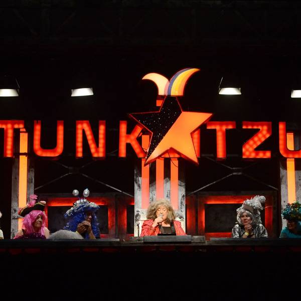 Biggi Wanninger sitzt umringt von weiteren Stunkern unter dem leuchtenden Logo der Stunksitzung.