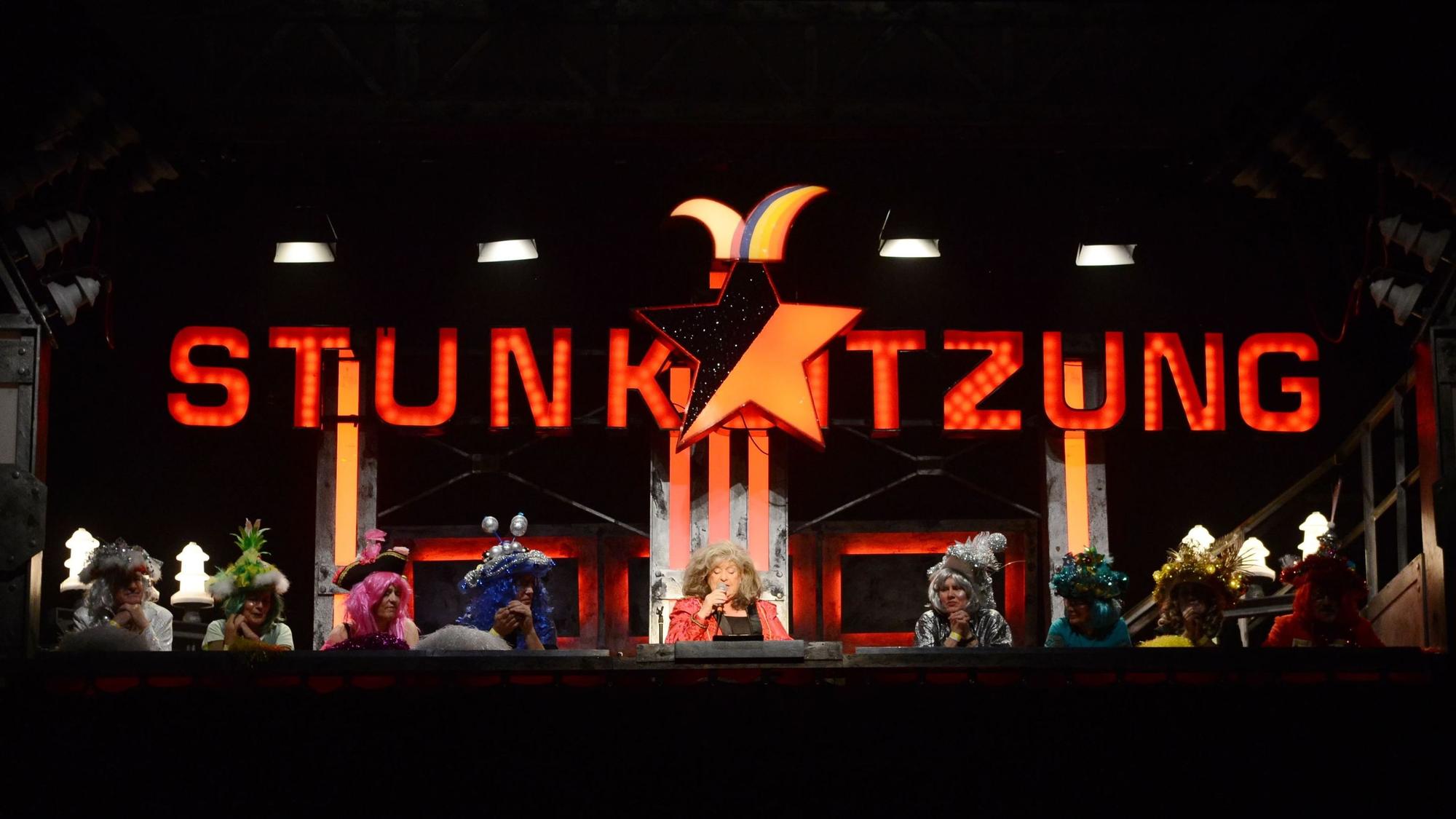 Biggi Wanninger sitzt umringt von weiteren Stunkern unter dem leuchtenden Logo der Stunksitzung.