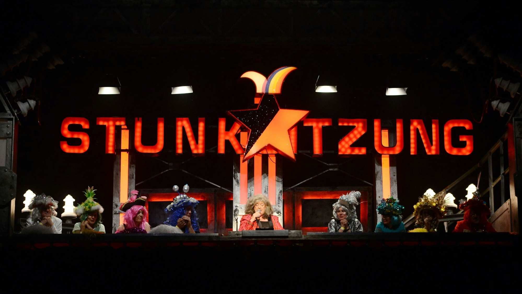 Biggi Wanninger sitzt umringt von weiteren Stunkern unter dem leuchtenden Logo der Stunksitzung.