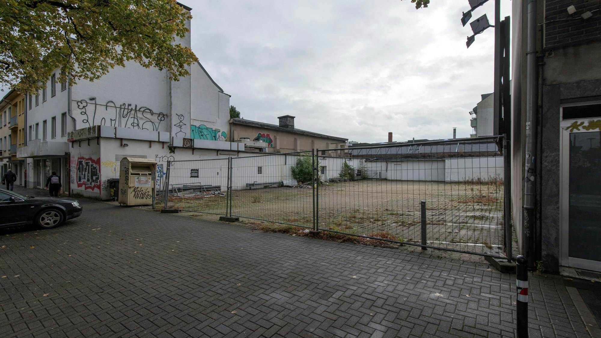 Hinter einem Bauzaun liegt eine Brachfläche, links und rechts sind Häuser.