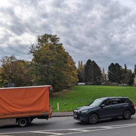 Autos und Transporter fahren an einer Wiese über eine stark befahrene Straße.