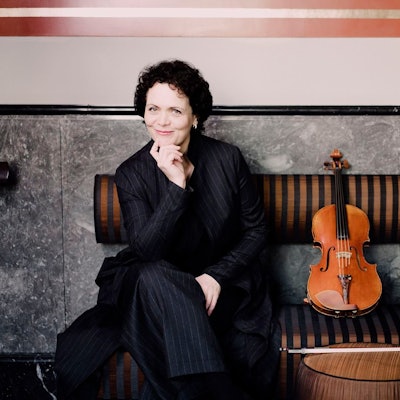 Tabea Zimmermann sitzt auf einer Bank und stützt ihr Kinn mit der Hand ab. Sie lächelt. Neben ihr auf der Bank ihre Bratsche.