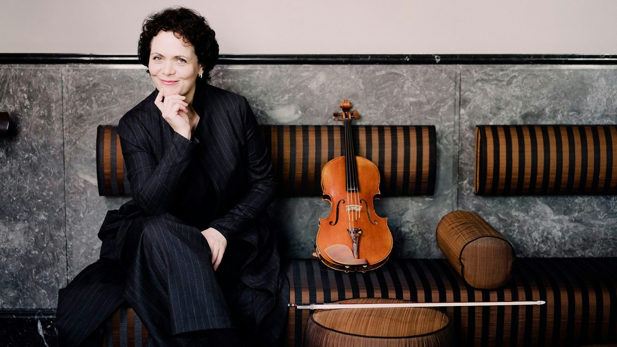 Tabea Zimmermann sitzt auf einer Bank und stützt ihr Kinn mit der Hand ab. Sie lächelt. Neben ihr auf der Bank ihre Bratsche.