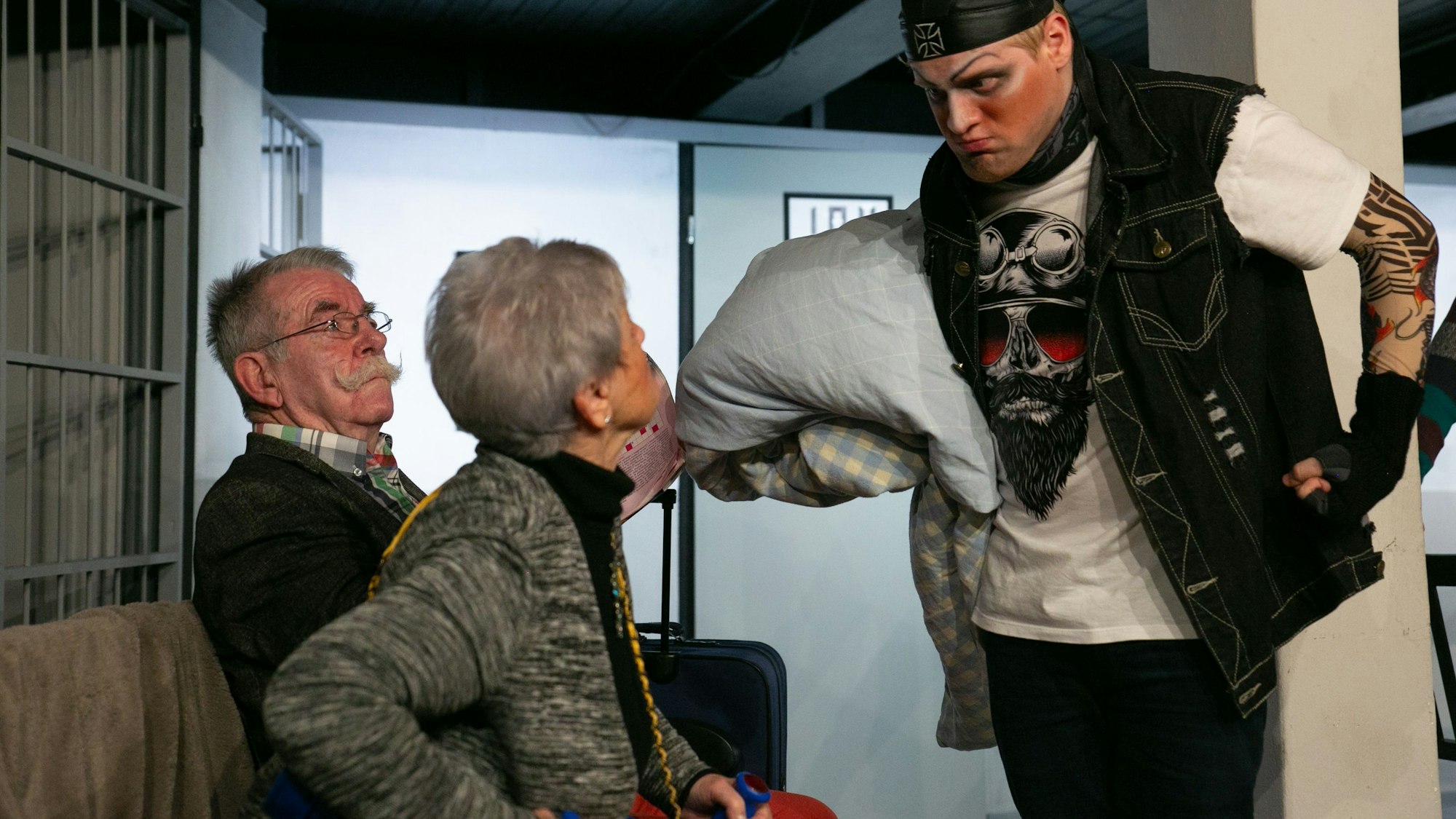 Ein junger Mann in Rocker-Kluft und bedrohlicher Pose steht vor einem älteren Paar.