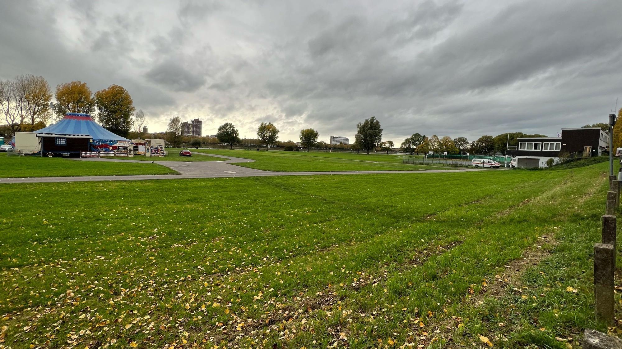 Dunkle Wolken hängen tief über einer großen Wiese am Rhein, im Hintergrund sind ein Zirkuszelt und ein Vereinsheim zu sehen.