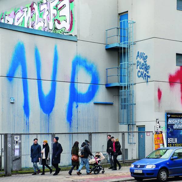 Menschen gehen auf einer Straße, an eine Häuserwand wurde zweimal „1 UP“ gesprüht.