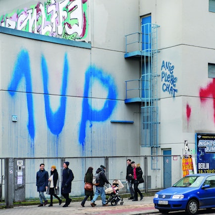 Menschen gehen auf einer Straße, an eine Häuserwand wurde zweimal „1 UP“ gesprüht.