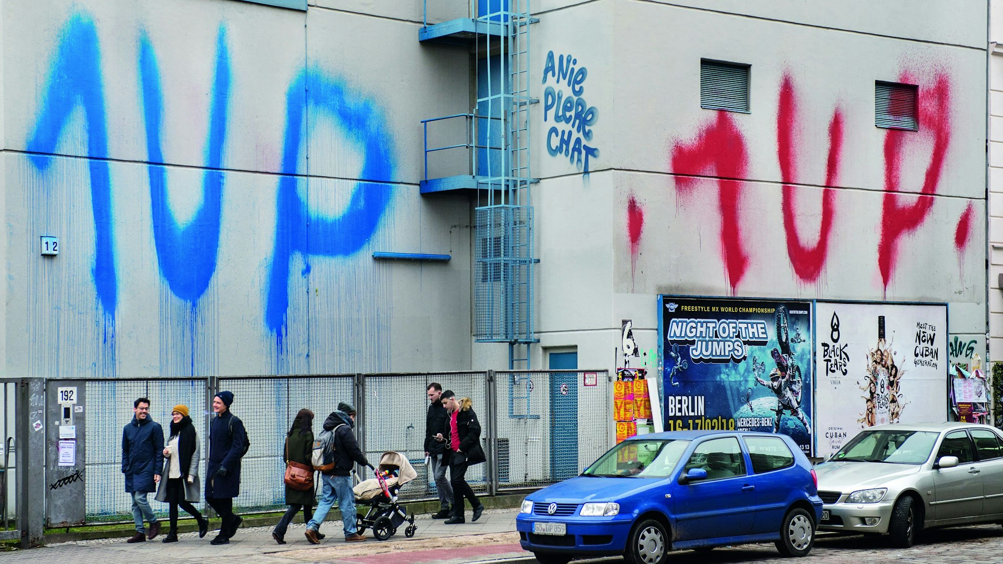 Menschen gehen auf einer Straße, an eine Häuserwand wurde zweimal „1 UP“ gesprüht.