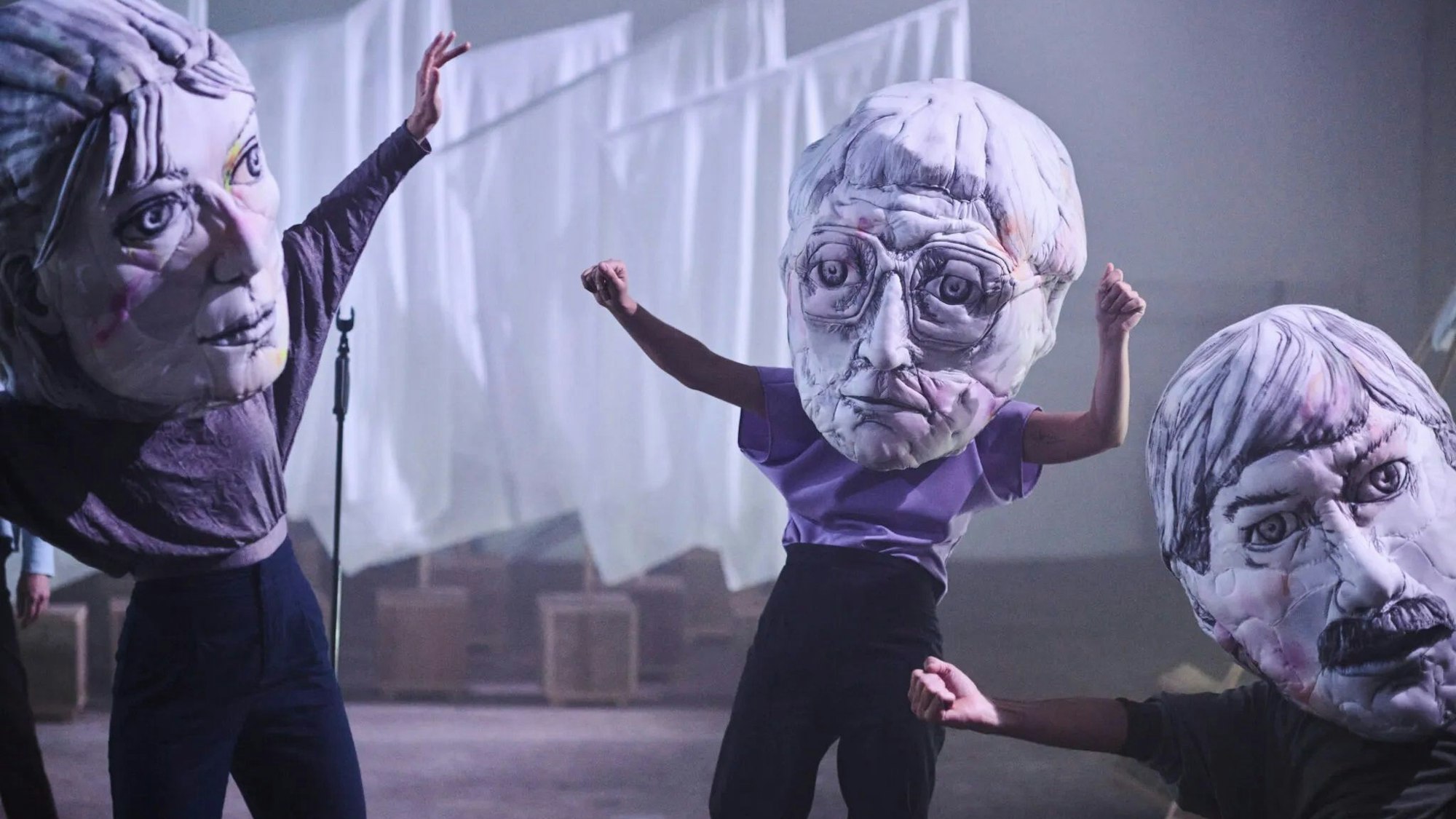 Auf dem Bild sieht man Schauspielende, die große Pappmascheeköpfe tragen. Die drei Darsteller haben die Arme in einer dynamischen Geste ausgestreckt, zwei von ihnen ballen dabei die Hände zu Fäusten.