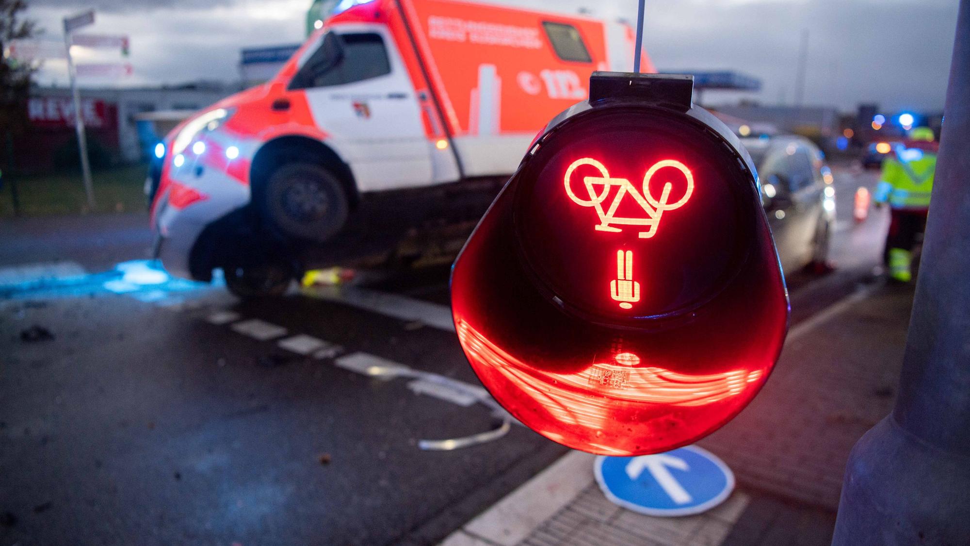 Das Symbolfoto zeigt eine bei einem Unfall beschädigte Ampel, deren rotes Leuchtzeichen auf dem Kopf hängt. Im Hintergrund ist ein beschädigter Rettungswagen zu sehen.
