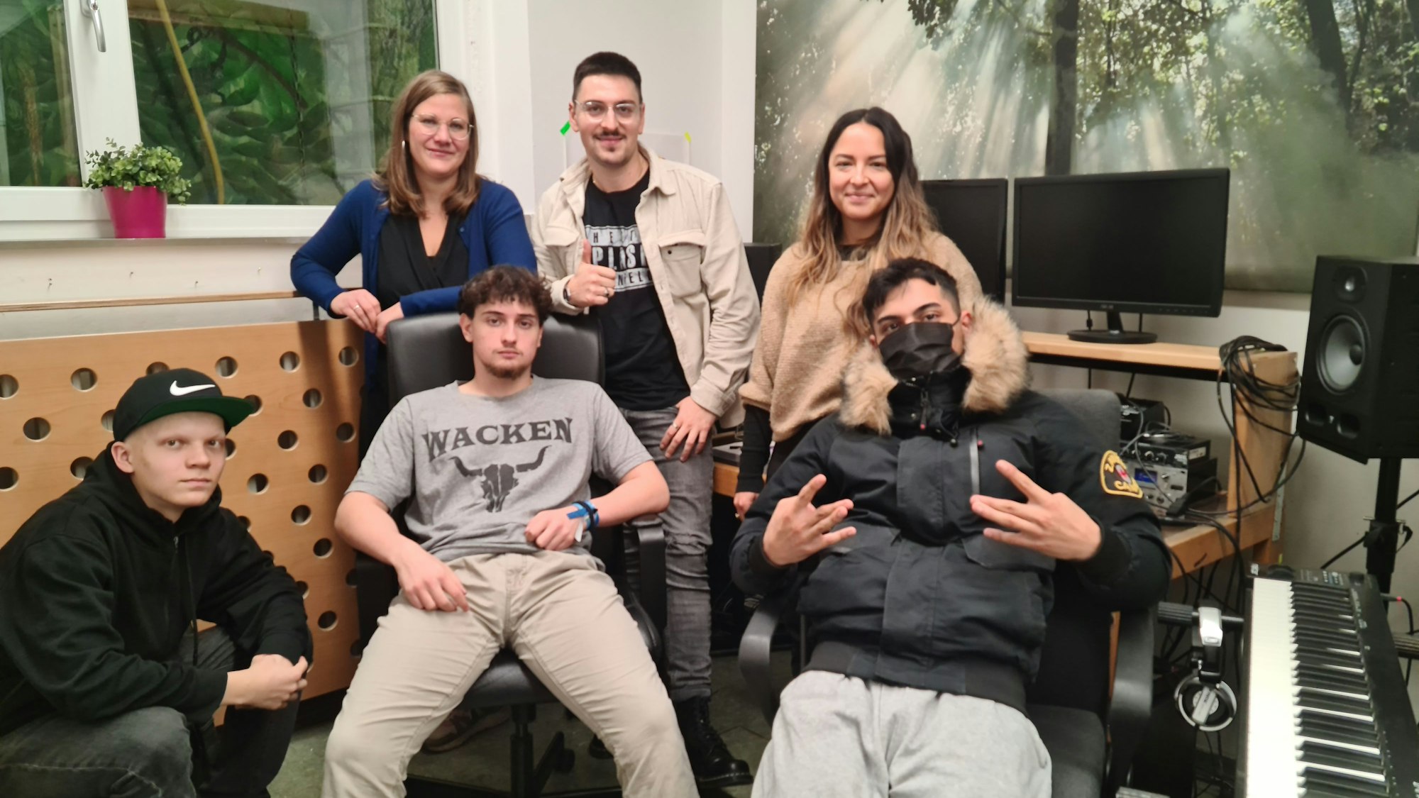 Vier junge Männer und zwei Frauen stehen und sitzen in einem Tonstudio.