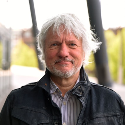 Kabarettist Jürgen Becker steht vor der Lanxess-Arena. Er trägt eine dunkle Jacke und blickt in die Kamera.