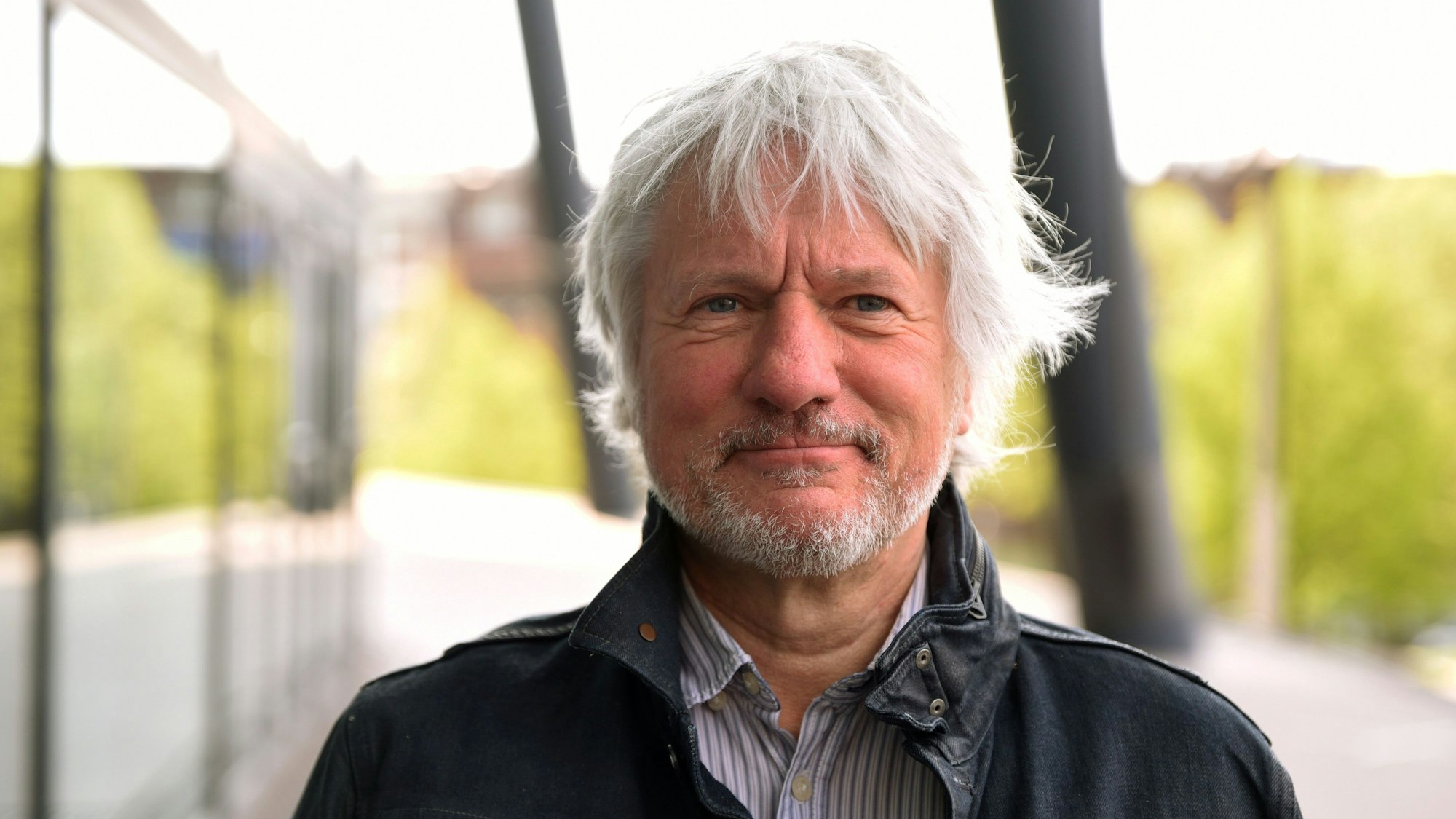 Kabarettist Jürgen Becker steht vor der Lanxess-Arena. Er trägt eine dunkle Jacke und blickt in die Kamera.