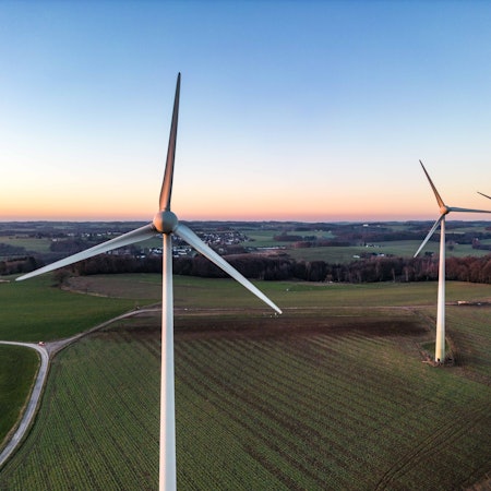 Drei große Windräder stehen auf einem Feld, schmale Straßen und ein Waldgebiet rahmen das Feld ein, im Hintergrund liegt eine kleine Siedlung.