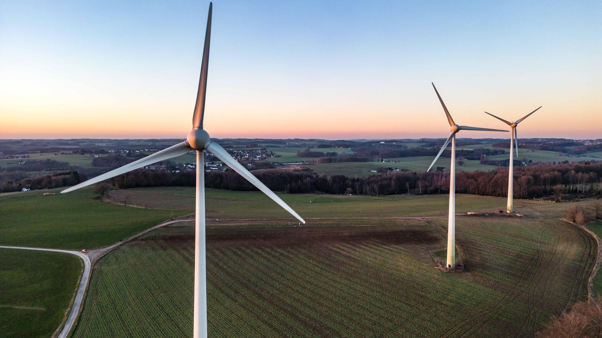 Drei große Windräder stehen auf einem Feld, schmale Straßen und ein Waldgebiet rahmen das Feld ein, im Hintergrund liegt eine kleine Siedlung.