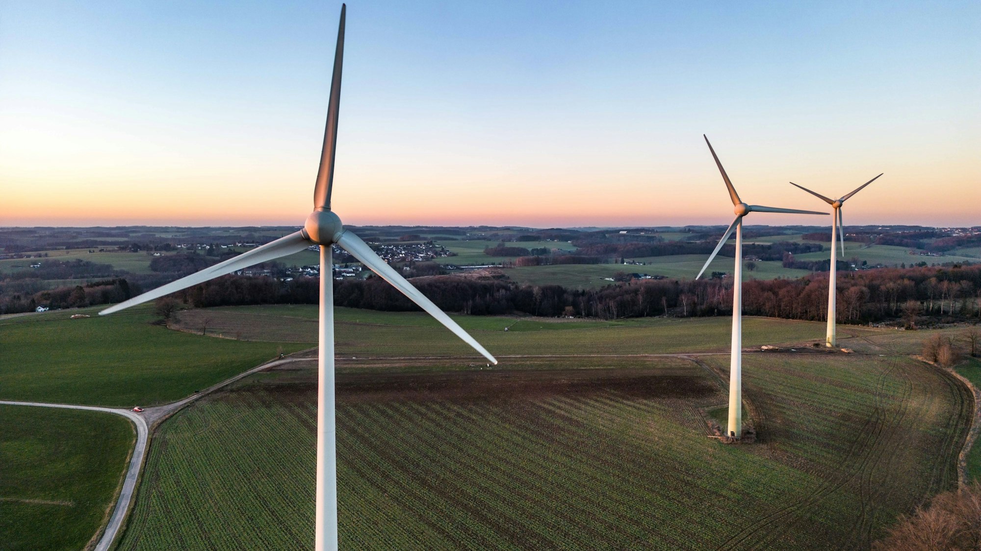 Drei große Windräder stehen auf einem Feld, schmale Straßen und ein Waldgebiet rahmen das Feld ein, im Hintergrund liegt eine kleine Siedlung.