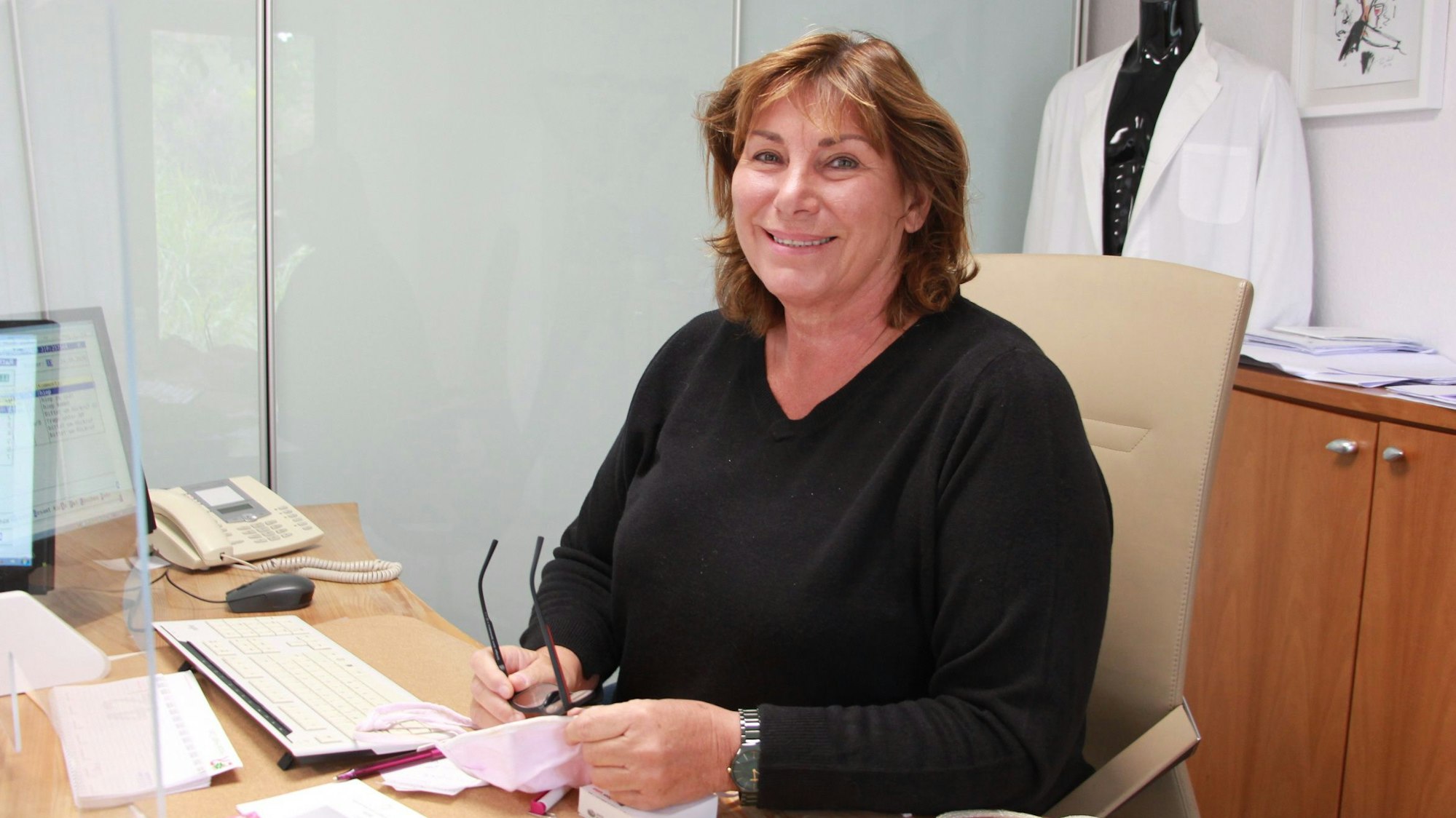 Eine Frau im schwarzen Pullover sitzt am Schreibtisch in einer Arztpraxis. Am Garderobenständer hängt ein weißer Kittel.