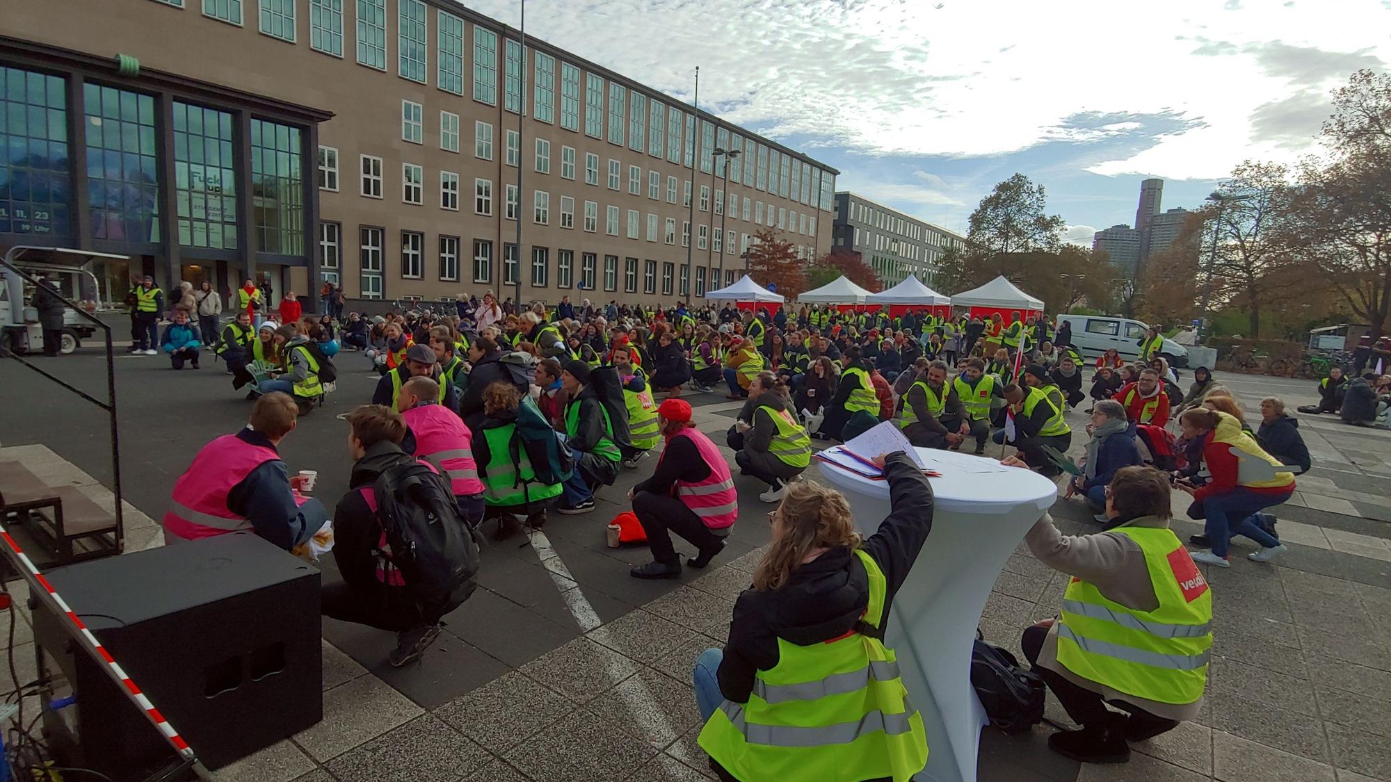 Viele Menschen in neongelben und -pinken Warnwesten, die auf einem Pltz in der Hocke sitzen.