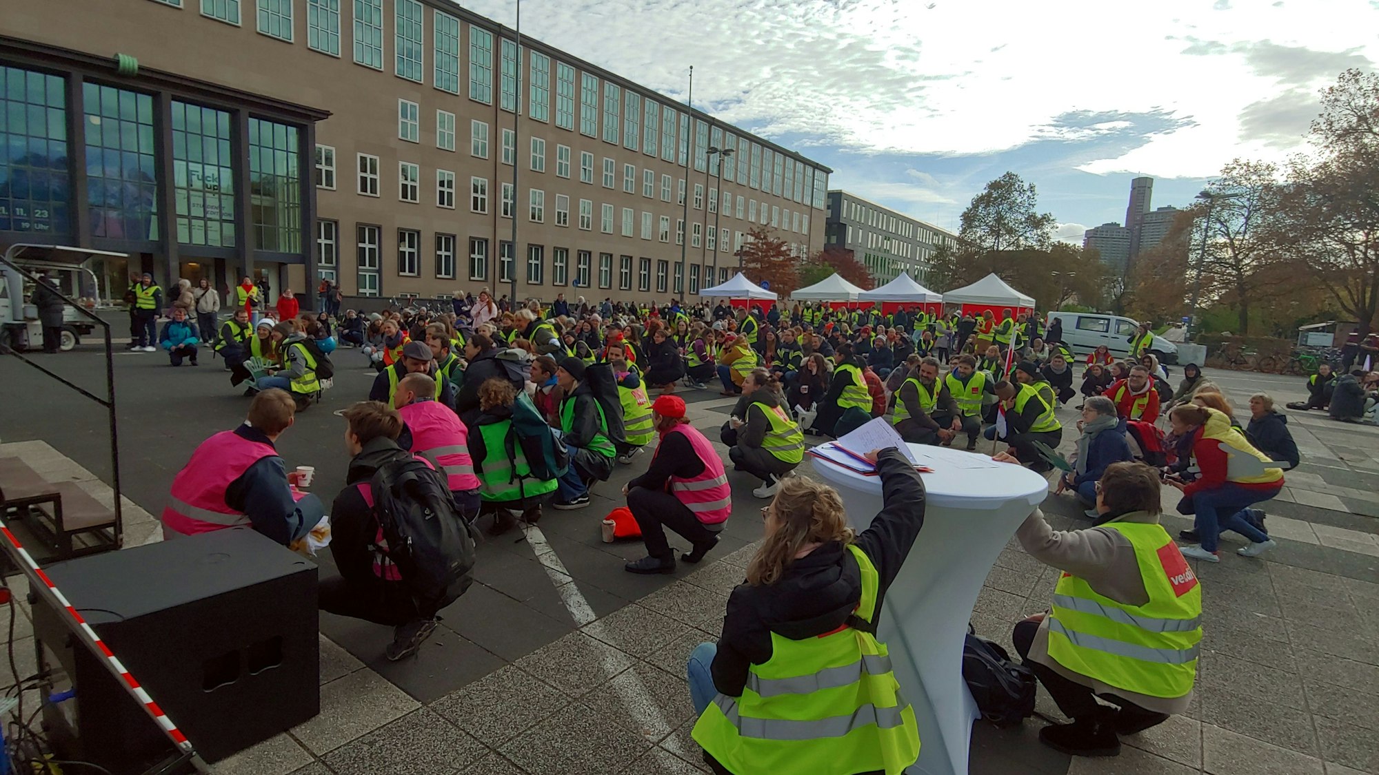 Viele Menschen in neongelben und -pinken Warnwesten, die auf einem Pltz in der Hocke sitzen.
