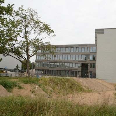 Ein modernes Bürogebäude mit vier Etagen; am Querbau markiert eine graue Fläche den geplanten Hotel-Anbau.