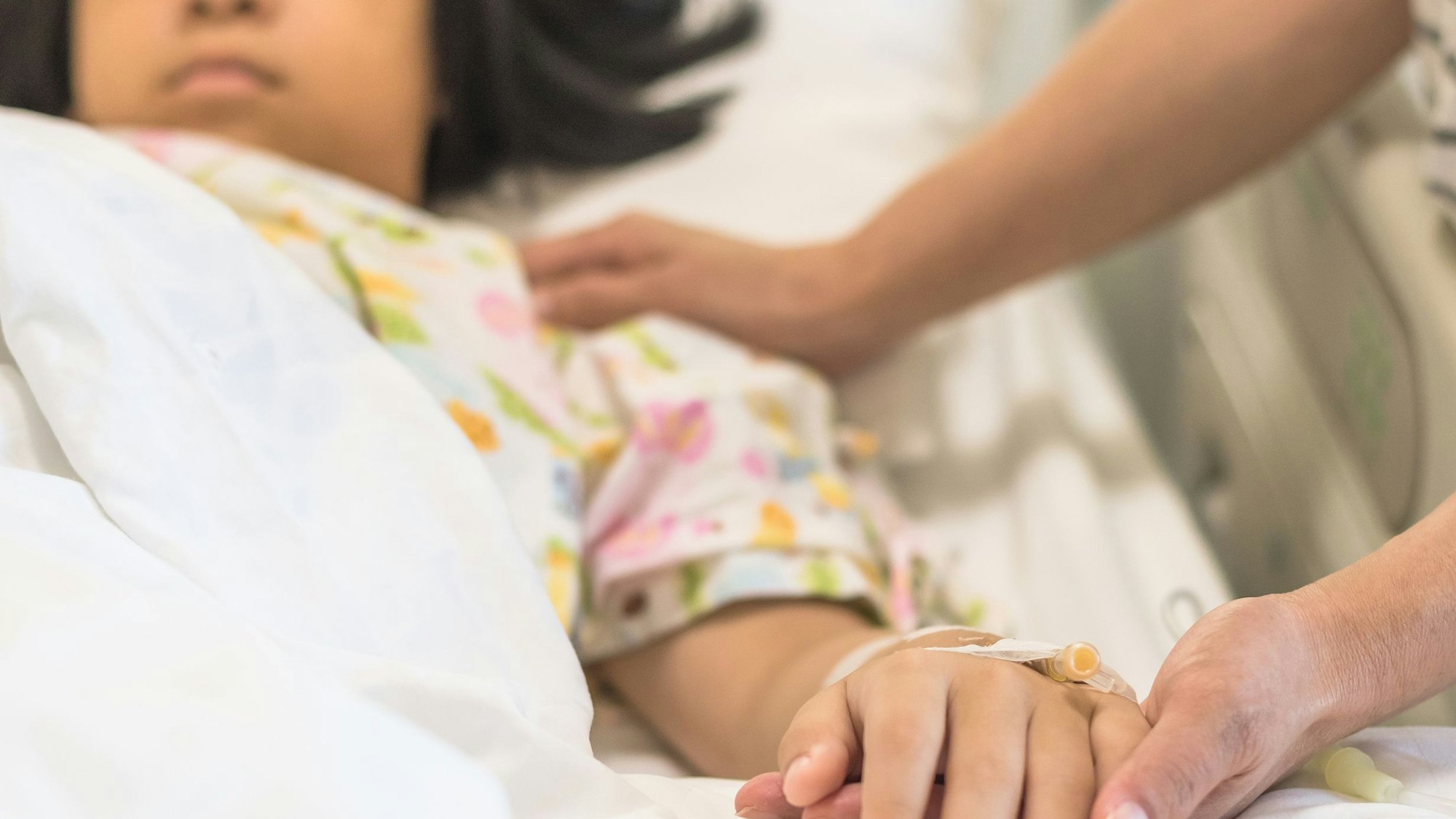 Ein Mädchen liegt im Bett, ein Erwachsener hält seine Hand, an der eine Infusionsnadel zu sehen ist.