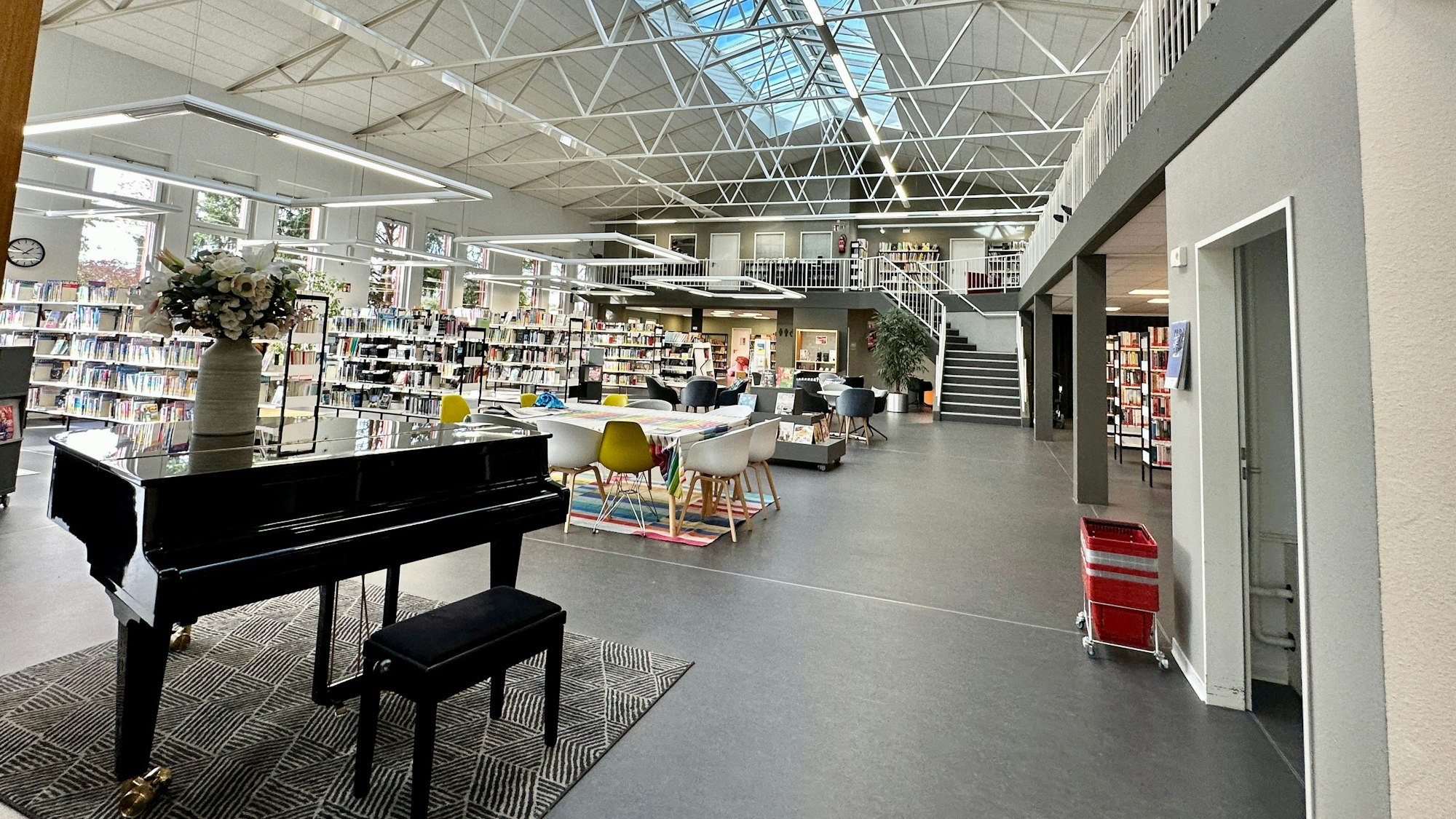 Ein hoher luftiger Raum mit einem Klavier im Vordergrund und Bücherregalen und einer Tischgruppe mit Stühlen ist zu sehen.