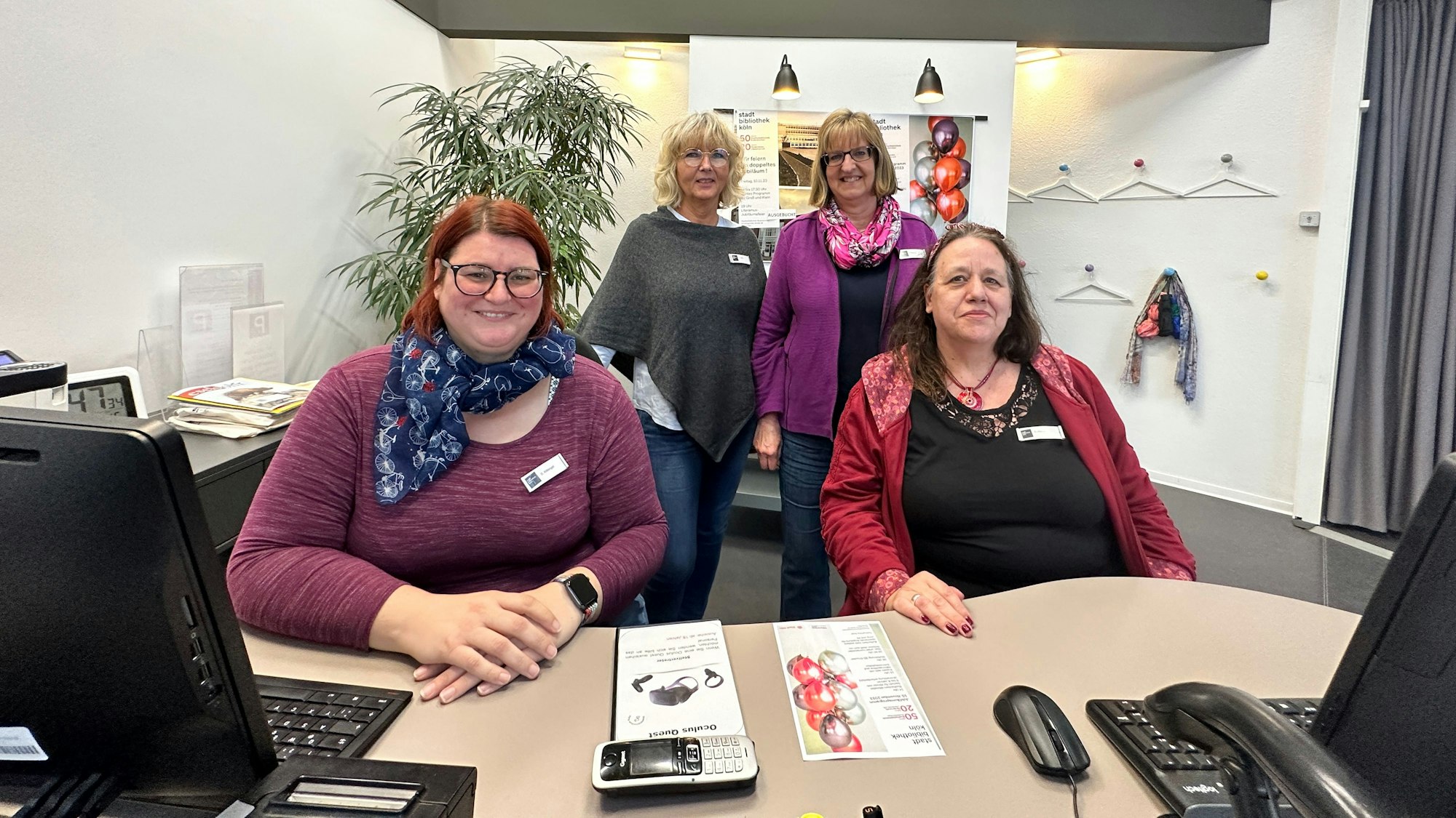Zwei Frauen stehen, zwei sitzen hinter einem Schreibtisch.
