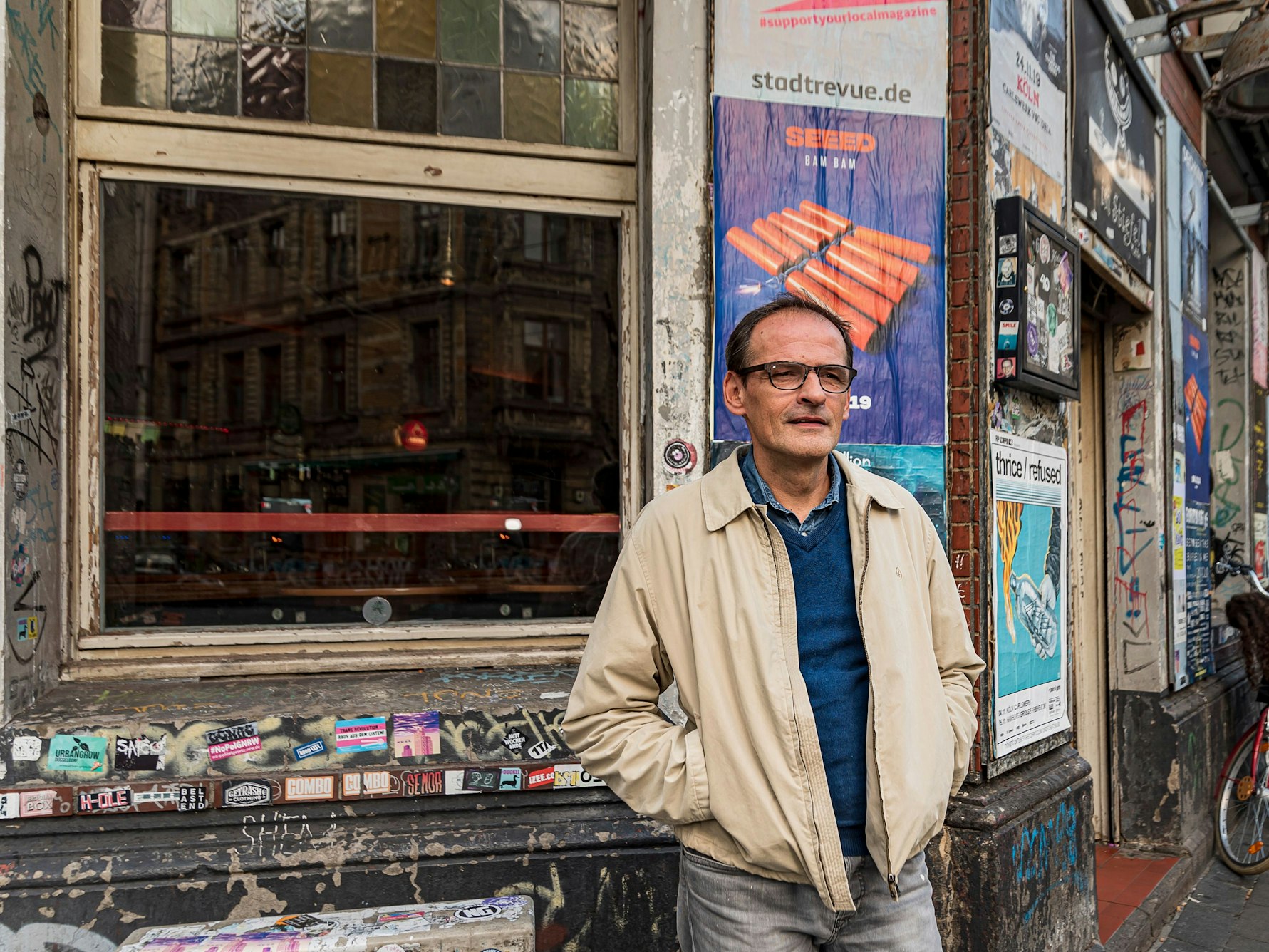 Stephan Freund steht vor der Fassade des Stiefels an der Zülpicher Straße. Die Fassade ist ziemlich heruntergerockt und mit Stickern und Plakaten beklebt