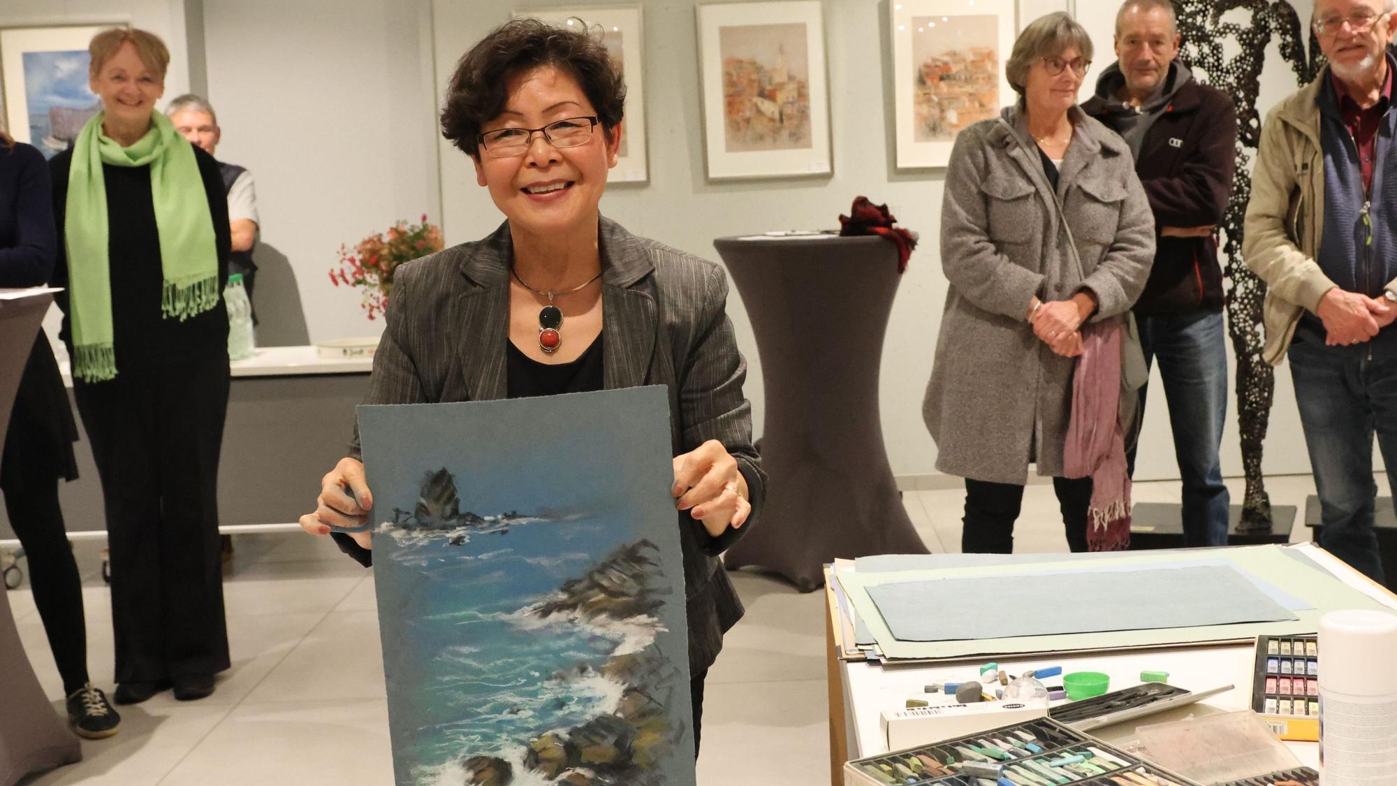 Künstlerin Hélène Michel hält eines ihrer Bilder hoch. Darauf sind Meereswellen zu sehen, die an eine Bucht schwappen. Im Hintergrund stehen Gäste in einer Ausstellung.