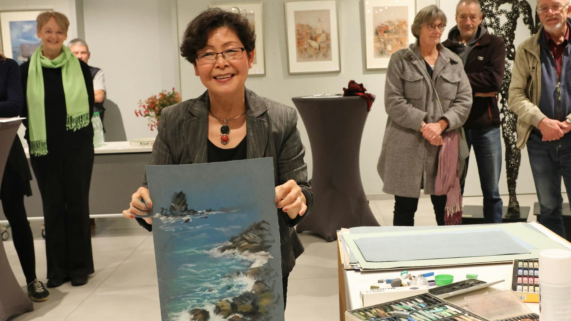 Künstlerin Hélène Michel hält eines ihrer Bilder hoch. Darauf sind Meereswellen zu sehen, die an eine Bucht schwappen. Im Hintergrund stehen Gäste in einer Ausstellung.