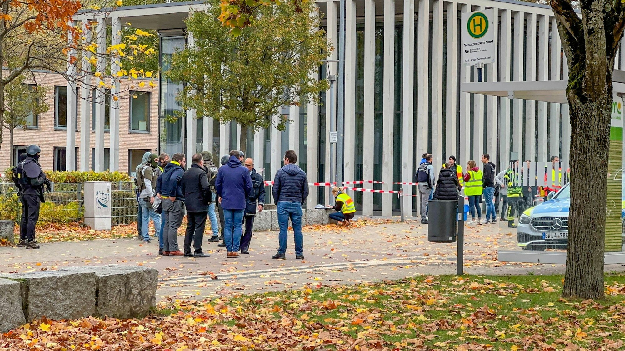 Menschen stehen vor einer Absperrung. Rechts sind eine Bushaltestelle und ein Polizeifahrzeug zu sehen.