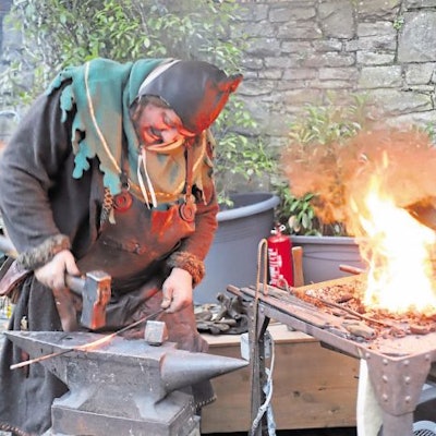 Ein Schmied in mittelalterlichem Kostüm bearbeitet mit einem schweren Hammer ein glühendes Rundeisen auf einem Amboss. Daneben lodert die Esse.