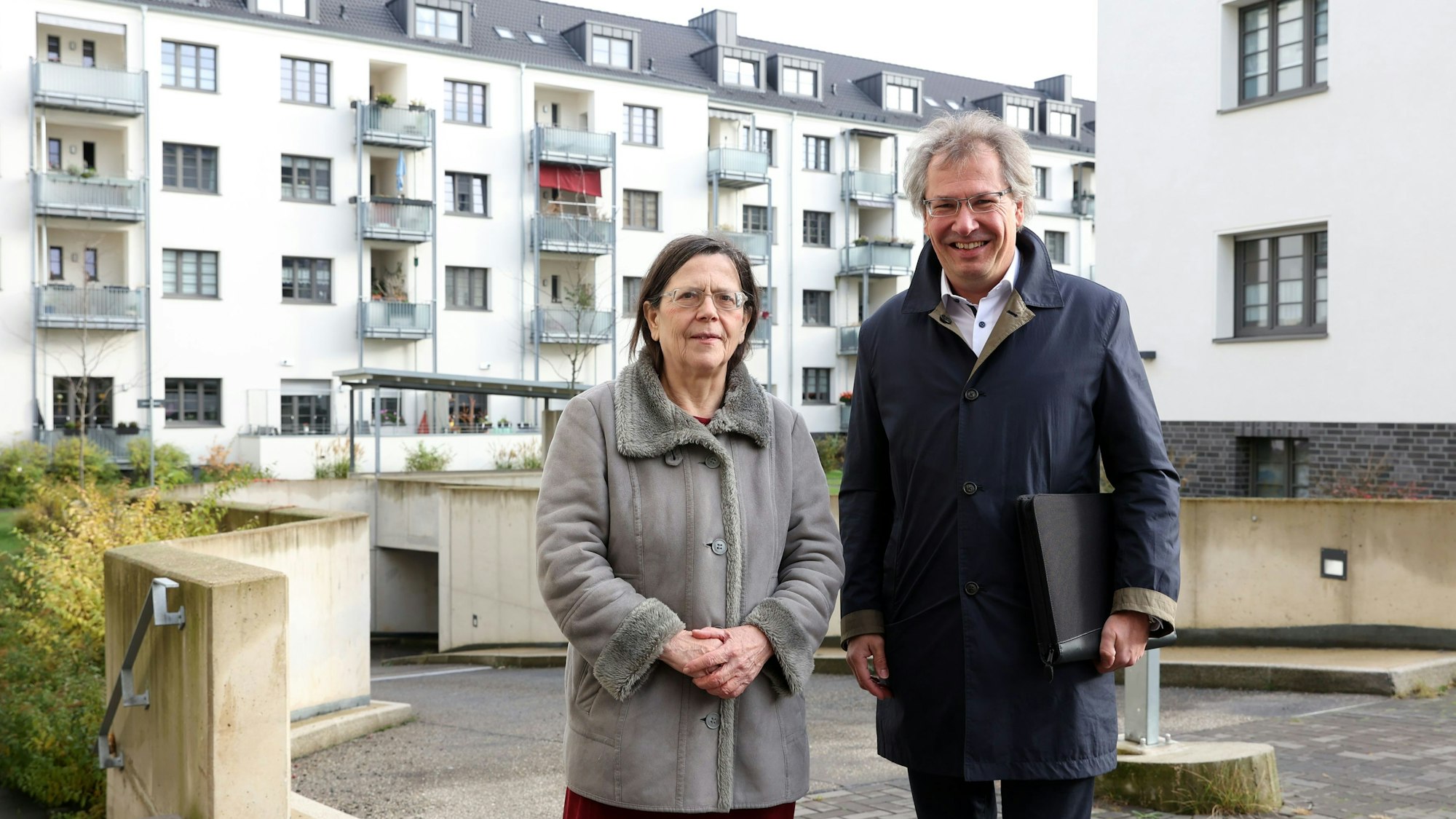 Eine Frau und ein Mann stehen vor mehreren Wohngebäuden und schauen in die Kamera.