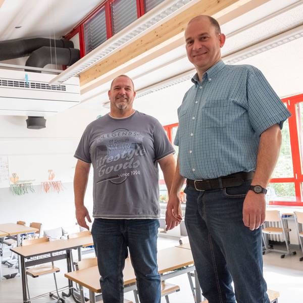 Christoph Breuer (v.l.), Frank Hecker und Andreas Kurth stehen in einem Klassenraum. Im Hintergrund ist ein Lüftungsgerät an der Decke zu sehen.
