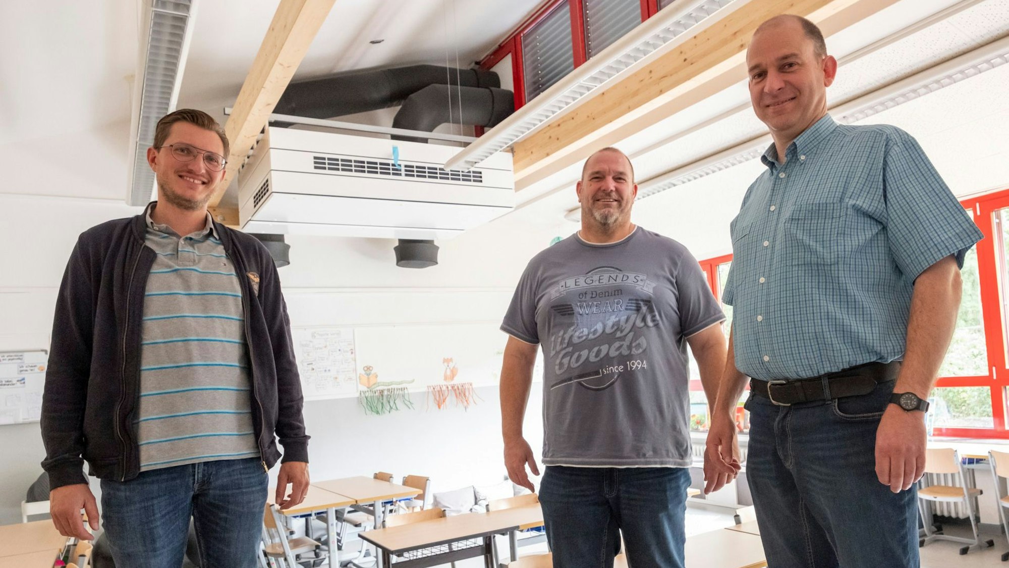 Christoph Breuer (v.l.), Frank Hecker und Andreas Kurth stehen in einem Klassenraum. Im Hintergrund ist ein Lüftungsgerät an der Decke zu sehen.