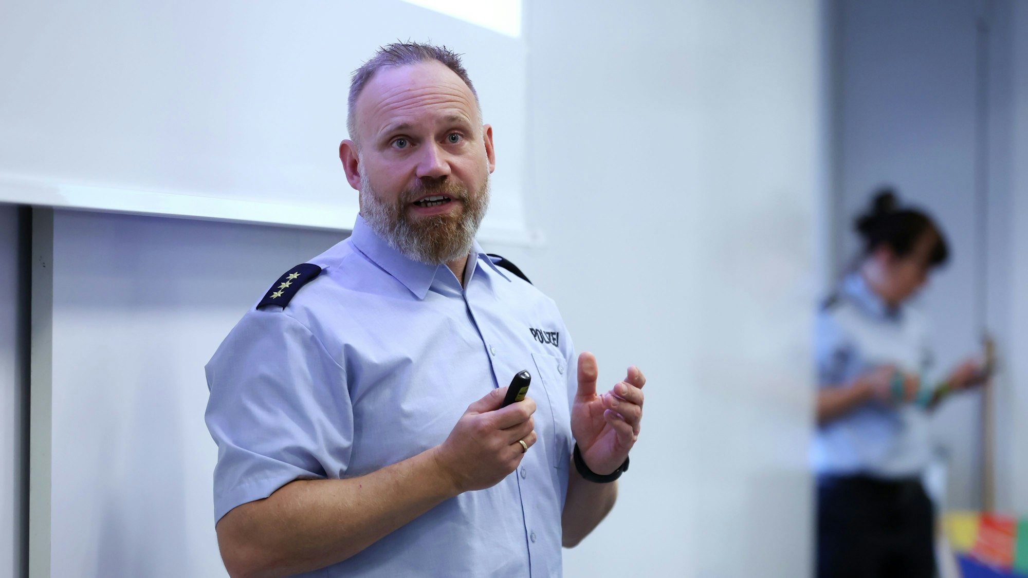Ein Polizist steht vor einer Leinwand, im Hintergrund ist eine Frau zu sehen.