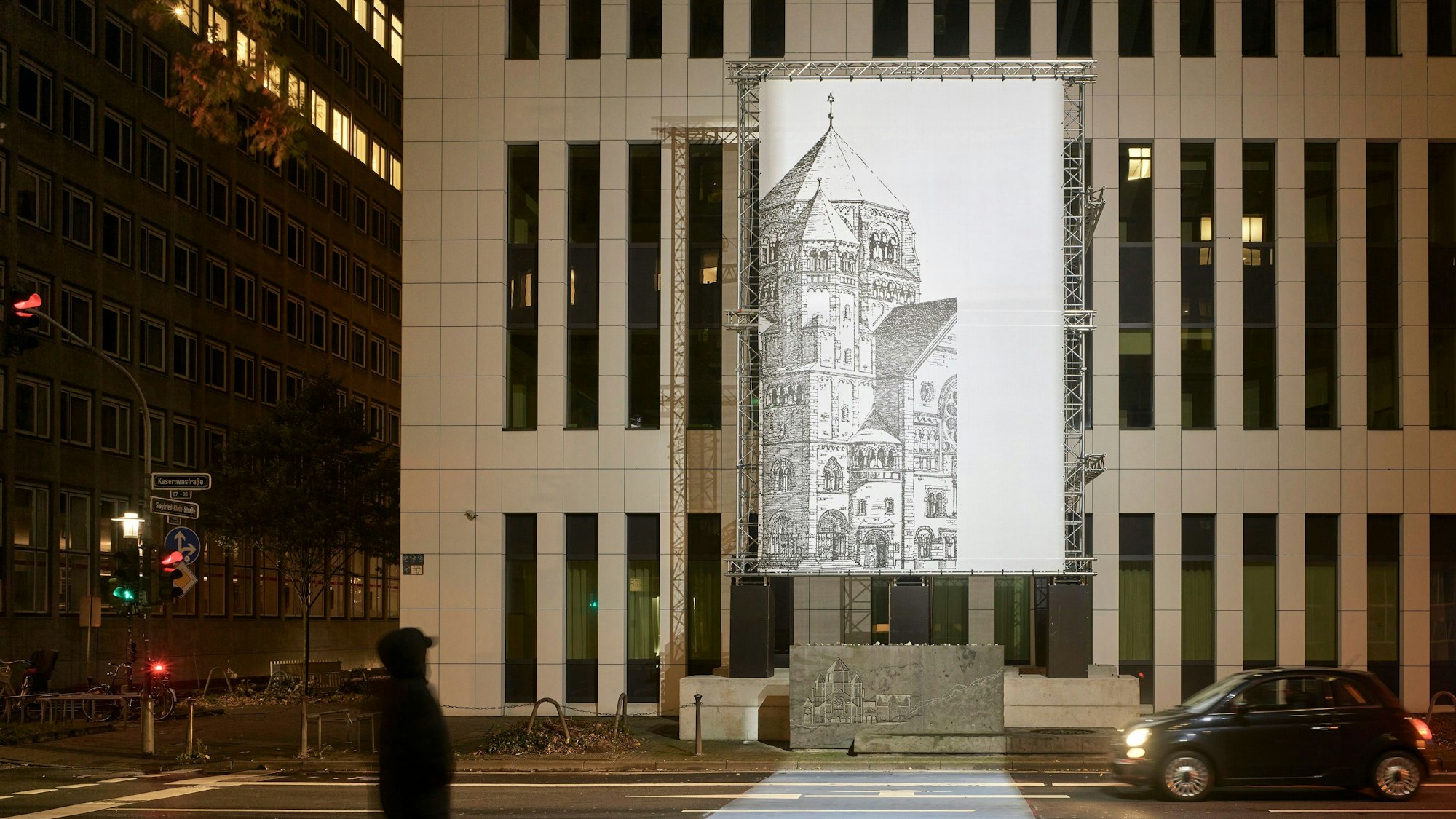 Das Bild zeigt über einem Gedenkstein und vor einem Hochhaus eine Installation. An Gerüsten hängt eine große Lichtinstallation, die den gezeichnet die zerstörte Synagoge in Düsseldorf zeigt.
