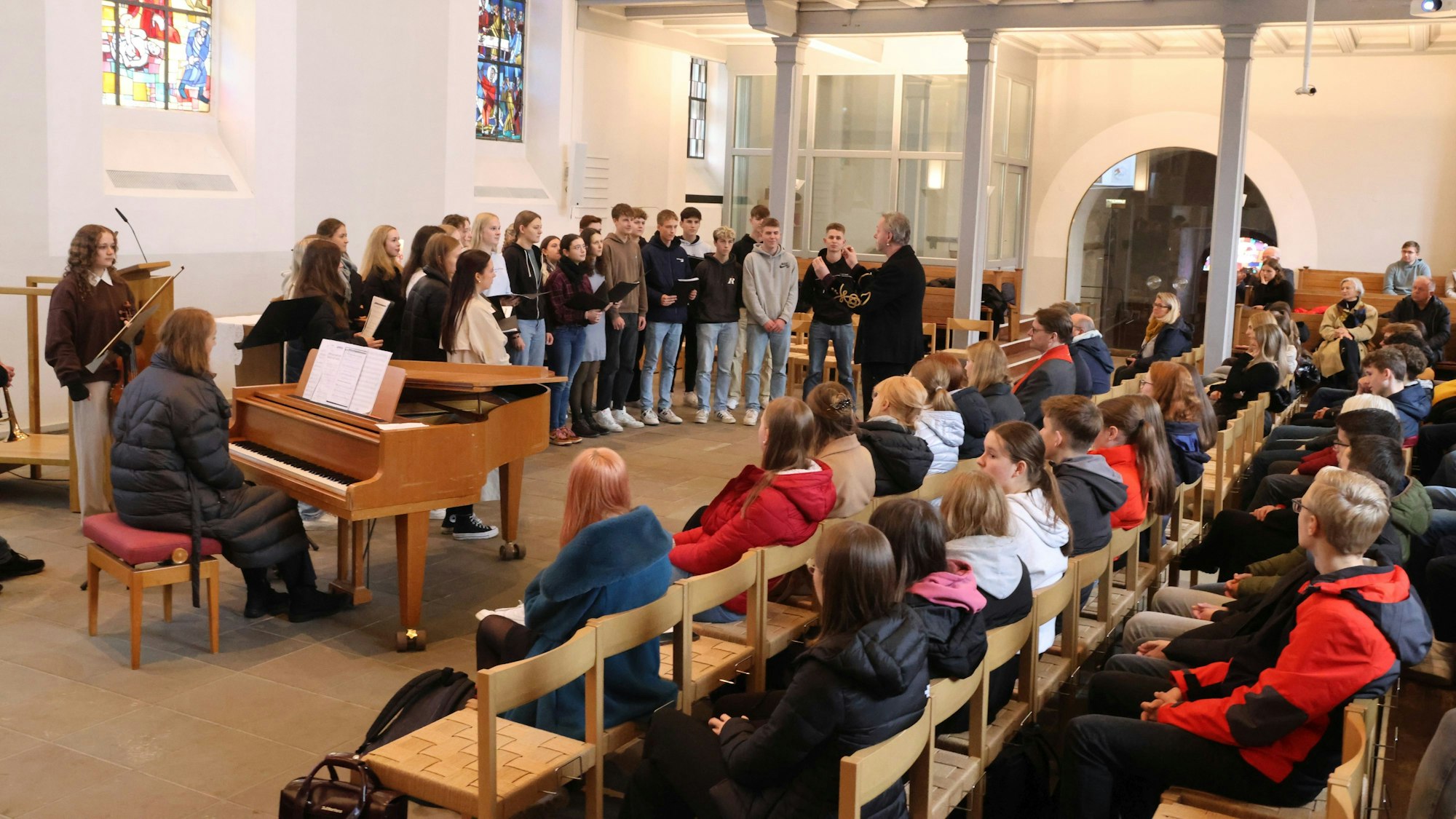 Eine Kirche ist voll besetzt. Im Altarraum steht eine Gruppe Schüler, eine Frau sitzt an einem Klavier.