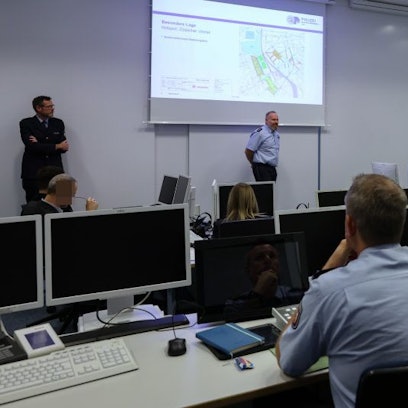 Frank Wißbaum, Einsatzleiter am 11.11, und Andreas Sandvoß, Leiter des Führungsstabs, stehen unter einer Leinwand und sprechen zu Kollegen.