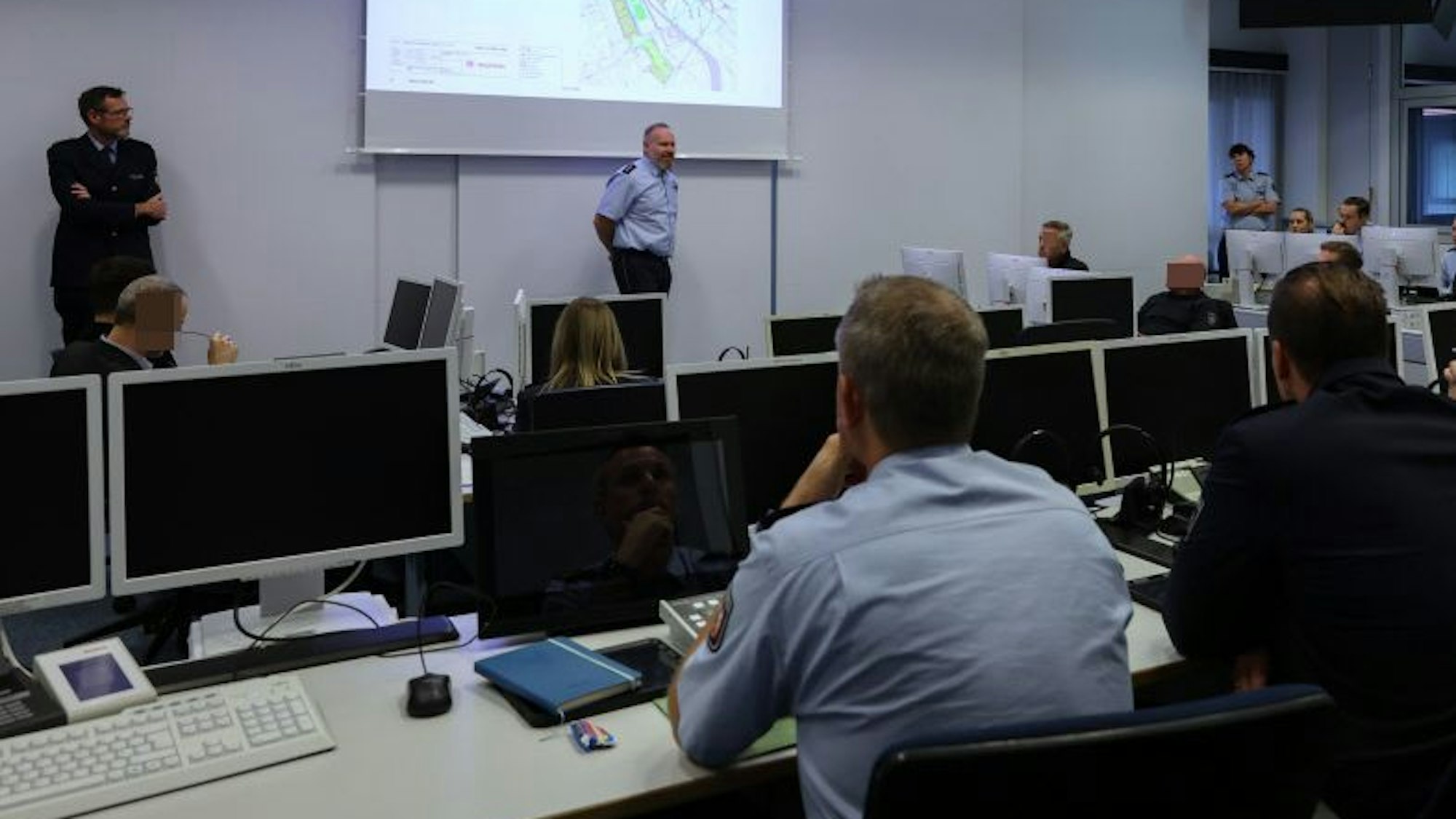 Frank Wißbaum, Einsatzleiter am 11.11, und Andreas Sandvoß, Leiter des Führungsstabs, stehen unter einer Leinwand und sprechen zu Kollegen.