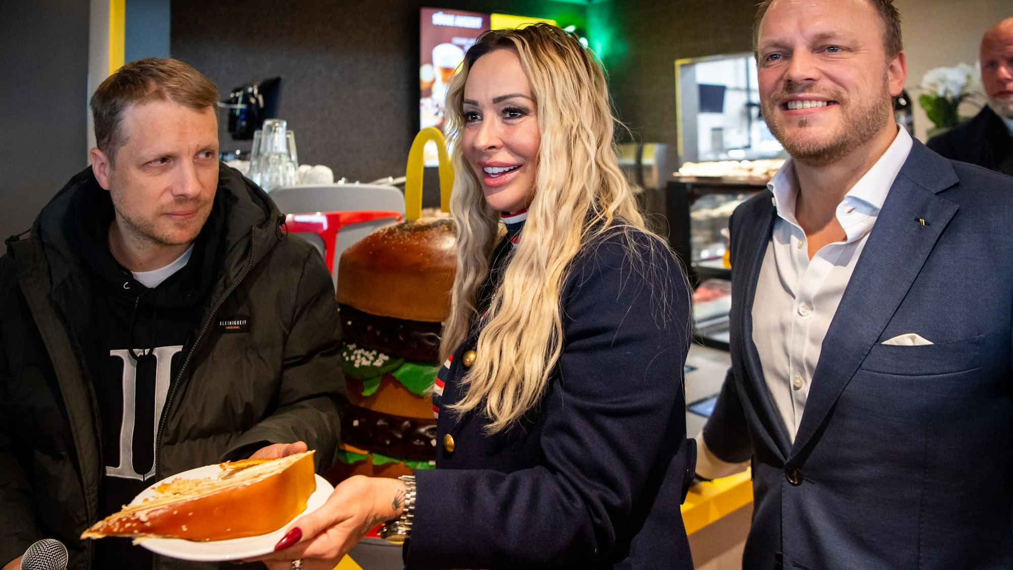 Oliver Pocher hält ein Mikrofon in der Hand, Cora Schumacher einen Teller mit einem Stück Kuchen. Daneben steht Christoph Mürtz.