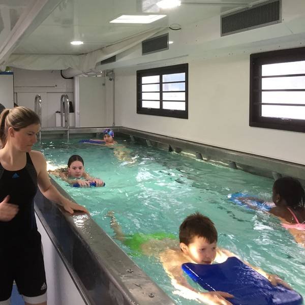 Kinder schwimmen in einem kleinen Becken in einem umgebauten Übersee-Container. Eine Schwimmlehrerin steht am Rand des Beckens.