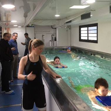 Kinder schwimmen in einem kleinen Becken in einem umgebauten Übersee-Container. Eine Schwimmlehrerin steht am Rand des Beckens.