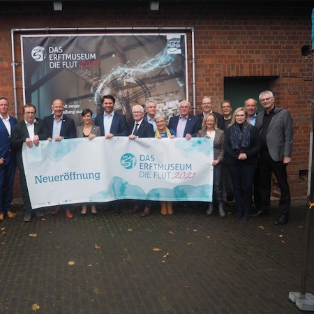 Eine Gruppe steht vor einem Plakat und hält ein Transparent, das auf die Eröffnung der Ausstellung hinweist.