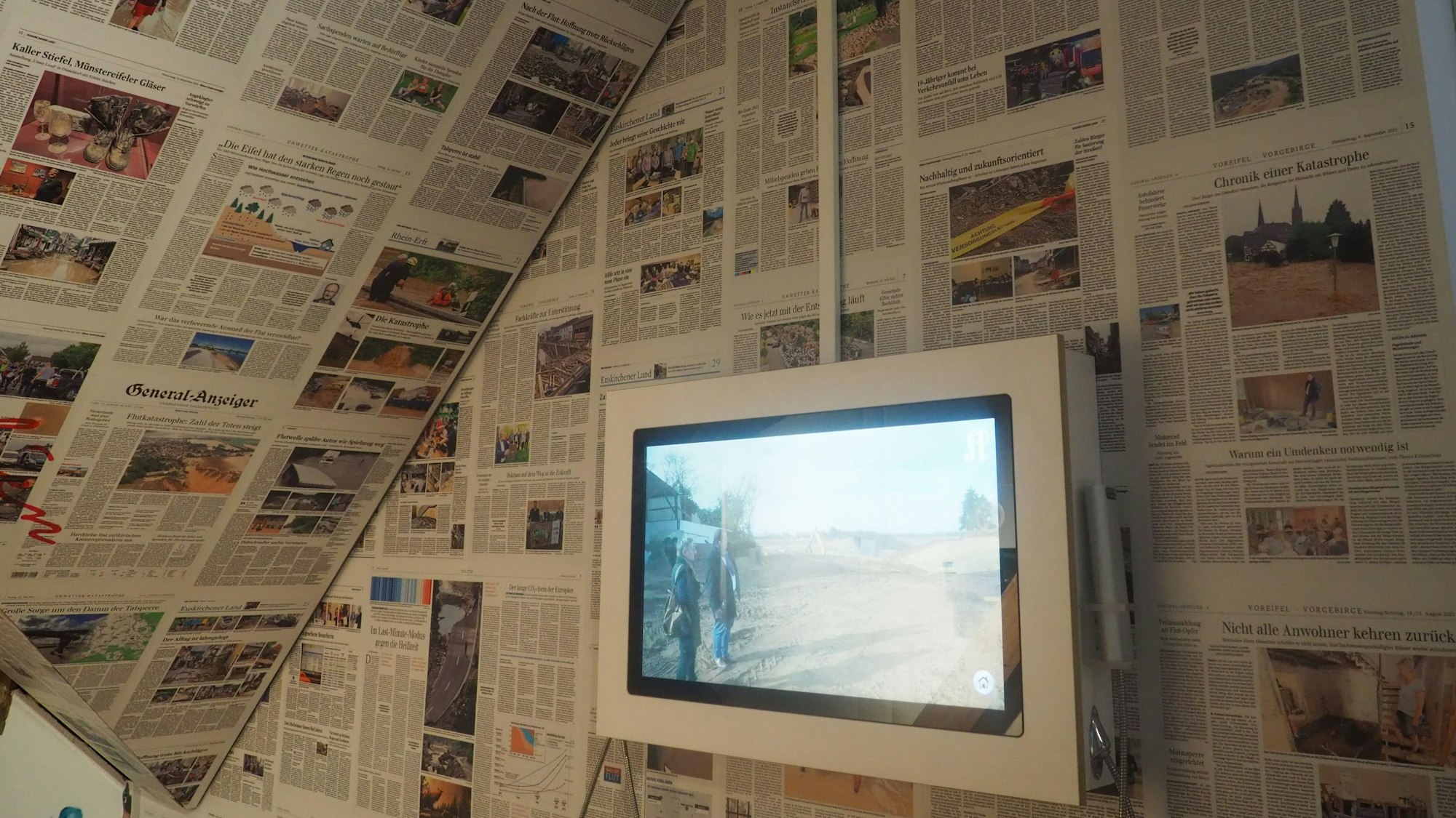 Decke und Wand sind mit Zeitungsseiten bedeckt, davor läuft auf einem Monitor ein Video
