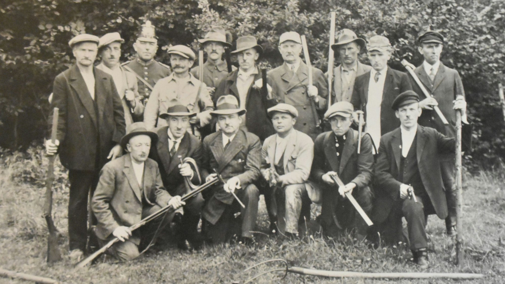 Mit einfachen Jagdgewehren und Geräten für die Feldarbeit posieren Männer auf einer Wiese.