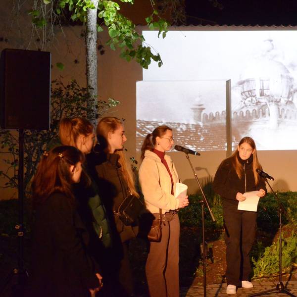 Schülerinnen an Mikrofonen tragen während der Gedenkfeier Texte vor, während ein auf eine Wand projiziertes Bild im Hintergrund die brennende Euskirchener Synagoge zeigt.