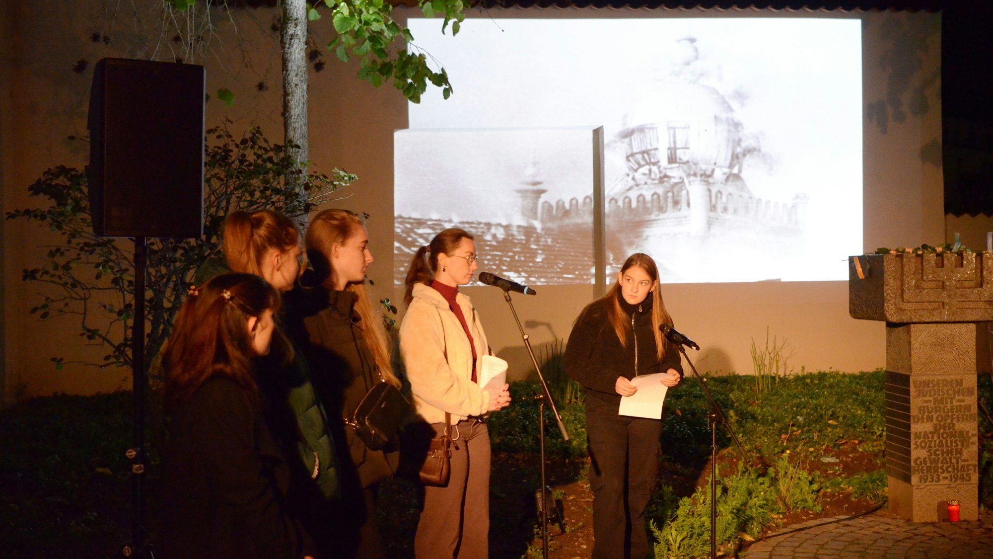 Schülerinnen an Mikrofonen tragen während der Gedenkfeier Texte vor, während ein auf eine Wand projiziertes Bild im Hintergrund die brennende Euskirchener Synagoge zeigt.